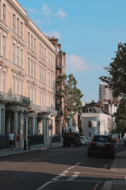 A street scene in Fulham featuring a row of elegant, cream-coloured Victorian-style townhouses with decorative cornices and tall windows, situated along a quiet road lined with trees and parked cars. In the foreground, a black van is positioned on the pavement, with part of its rear doors open, indicating recent loading or unloading. Nearby, boxes wrapped in plastic and cardboard, along with pieces of furniture such as a small table, are visible being carried or prepared for transport by Man and Van Fulham, a professional removals company. The image captures the furniture transport process of home relocation, with the partially visible interior of a property in the background, and the bright daylight providing clear visibility of the moving activities. Overall, the scene exemplifies the logistical planning involved in residential removals, with furniture and packing materials being carefully loaded onto the van for delivery at the new property.