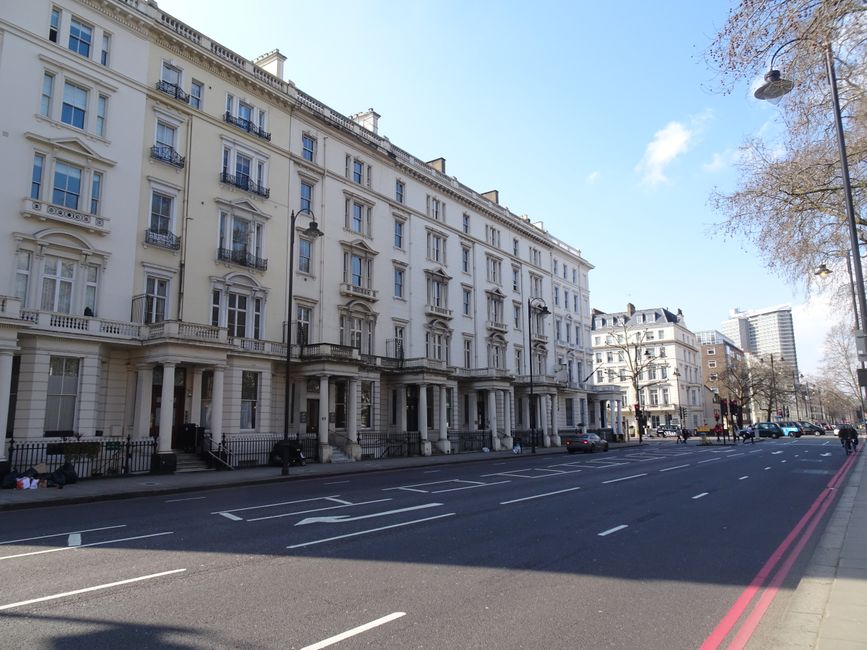 A wide street scene featuring a large, multi-storey white residential building with classic architectural details, including decorative window moldings, small balconies with black wrought iron railings, and columned porch entrances at ground level. The building is situated on the left side of the image, with a clear blue sky overhead and patches of sunlight illuminating the facade. The street is relatively empty, with a few parked cars along the curb and some pedestrians walking on the pavement. On the right side, there are leafless trees, street lamps, and modern high-rise buildings visible in the background. The pavement area next to the building appears clear, with some cardboard boxes and plastic wrapping on the sidewalk. The scene suggests a calm urban environment suitable for house removals or furniture transport, with street access conducive to loading and unloading items, supporting reliable moving services by Man and Van Fulham.
