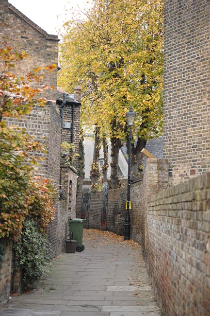 A narrow residential alleyway with a paved stone surface, lined by brick walls on both sides, leading to a small staircase. Tall trees with yellow and orange autumn leaves overhang the alley, partially covering the view of houses and buildings in the background. A black street lamp is mounted on a pole along the path, and a green waste bin is positioned near the left wall. This scene illustrates a typical backstreet in Fulham, with potential challenges for home relocation and furniture transport along narrow streets. Man and Van Fulham, a professional removals service, may assist with packing and loading during house removals in such confined urban environments.